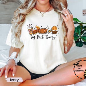 Puede incluir: Camiseta de color marfil con un gr&aacute;fico de alimentos de oto&ntilde;o, que incluye un caf&eacute; con leche de calabaza especiada, tarta, pavo y magdalena. El texto "Big Back Energy" est&aacute; impreso debajo. Un gr&aacute;fico de murci&eacute;lago y casa embrujada est&aacute; en la parte inferior derecha.