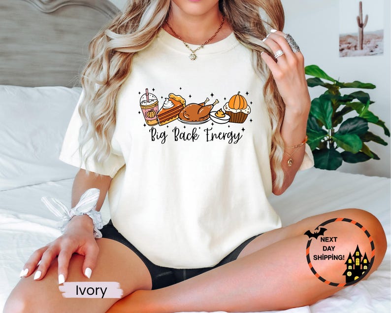 Puede incluir: Camiseta de color marfil con un gr&aacute;fico de alimentos de oto&ntilde;o, que incluye un caf&eacute; con leche de calabaza especiada, tarta, pavo y magdalena. El texto "Big Back Energy" est&aacute; impreso debajo. Un gr&aacute;fico de murci&eacute;lago y casa embrujada est&aacute; en la parte inferior derecha.