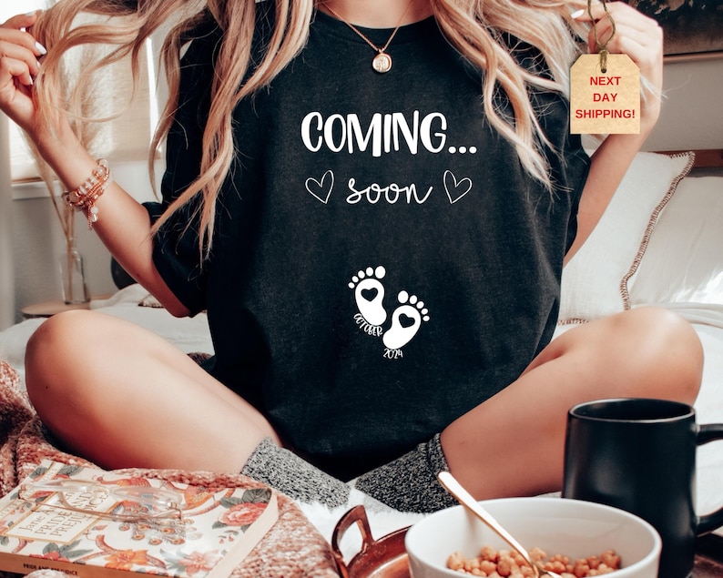Op de afbeelding: Een zwart t-shirt met witte tekst die COMING... soon leest, samen met twee kleine hartjes. Onder de tekst staan twee babyvoetjes met hartvormige hielen, met de woorden OCTOBER en 2024 onder de voetafdrukken gedrukt.