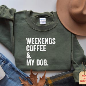 May include: An olive green sweatshirt with the text "WEEKENDS COFFEE & MY DOG" in white. The sweatshirt is displayed with a brown hat, a pair of jeans, and a coffee cup.