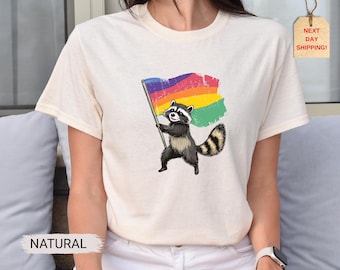 May include: Cream-colored t-shirt featuring a raccoon holding a rainbow flag. The raccoon is depicted in black, gray, and white, with a striped tail. The flag has a distressed, textured appearance. The word "NATURAL" is printed on the bottom left.
