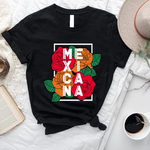 May include: Black t-shirt featuring the word "MEXICANA" in white letters, integrated with red, orange, and green floral graphics. The design is framed in a white rectangle. The shirt is displayed on a white surface with a hat and coffee cup.