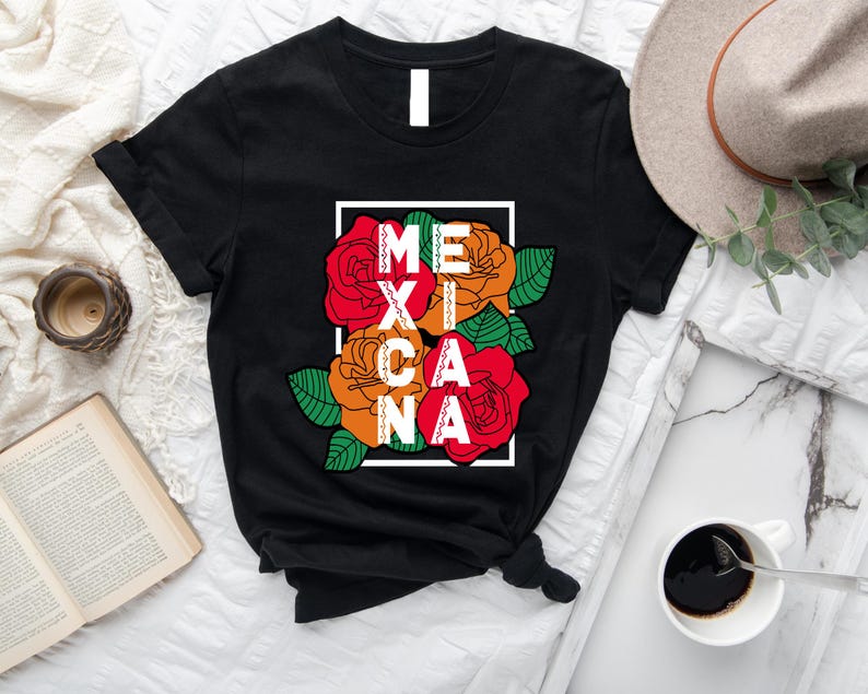 May include: Black t-shirt featuring the word "MEXICANA" in white letters, integrated with red, orange, and green floral graphics. The design is framed in a white rectangle. The shirt is displayed on a white surface with a hat and coffee cup.