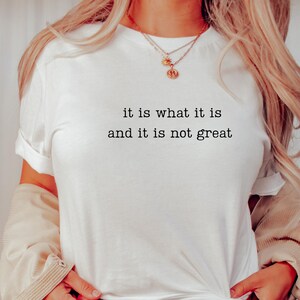 May include: White t-shirt with the text "it is what it is and it is not great" in a black, simple font. The shirt has rolled-up sleeves and is being worn with a beige jacket and gold necklaces.