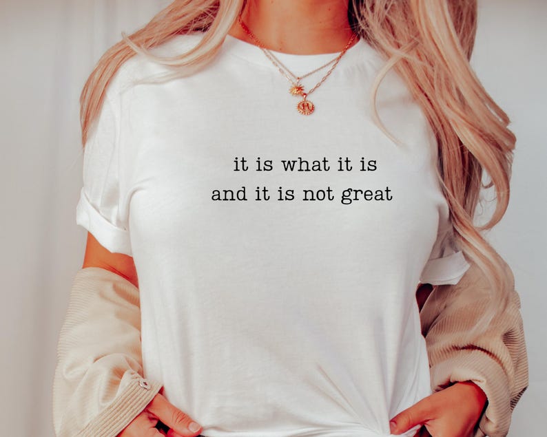 May include: White t-shirt with the text "it is what it is and it is not great" in a black, simple font. The shirt has rolled-up sleeves and is being worn with a beige jacket and gold necklaces.