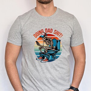 May include: A light gray t-shirt featuring a graphic of a skeleton on a toilet, using a phone. The graphic includes the text "DOING DAD SHIT!" in a circular design with a sunset background. The skeleton wears a "DAD" hat.