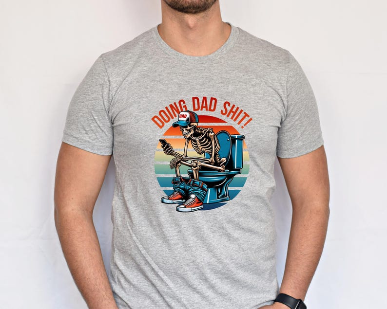 May include: A light gray t-shirt featuring a graphic of a skeleton on a toilet, using a phone. The graphic includes the text "DOING DAD SHIT!" in a circular design with a sunset background. The skeleton wears a "DAD" hat.