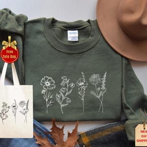 May include: An olive green sweatshirt with a white floral design. A beige tote bag with a matching floral print is included. A brown hat is placed on the top right. A tag says "Next Day Shipping!"