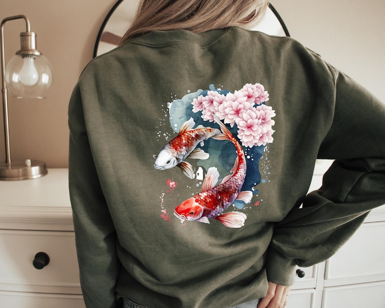 Puede incluir: Una sudadera verde con un dise&ntilde;o de acuarela de dos peces koi nadando entre flores de cerezo rosas.