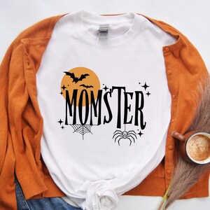 May include: White t-shirt with a black and orange graphic design that says "Momster" with a spider, bat, and moon. The shirt is paired with a brown cardigan.