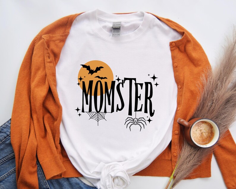 May include: White t-shirt with a black and orange graphic design that says "Momster" with a spider, bat, and moon. The shirt is paired with a brown cardigan.
