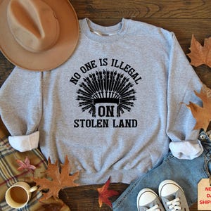 May include: A light gray sweatshirt with the text "NO ONE IS ILLEGAL ON STOLEN LAND" in black, featuring a Native American headdress design. The sweatshirt has white cuffs and is displayed with a hat, jeans, and fall leaves.