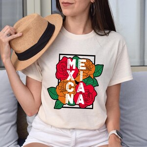 May include: Cream-colored t-shirt featuring the word "MEXICANA" in a black frame, with red and orange roses and green leaves. A straw hat with a black band is held in the hand.