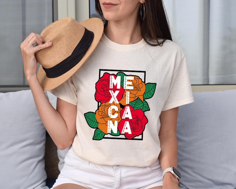 May include: Cream-colored t-shirt featuring the word "MEXICANA" in a black frame, with red and orange roses and green leaves. A straw hat with a black band is held in the hand.
