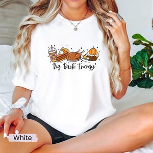 Puede incluir: Camiseta blanca con un gr&aacute;fico de comida de Acci&oacute;n de Gracias: un latte de especias de calabaza, tarta, pavo y una magdalena de calabaza. El texto "Big Pack Energy" est&aacute; escrito en negro. La camiseta es de estilo casual.