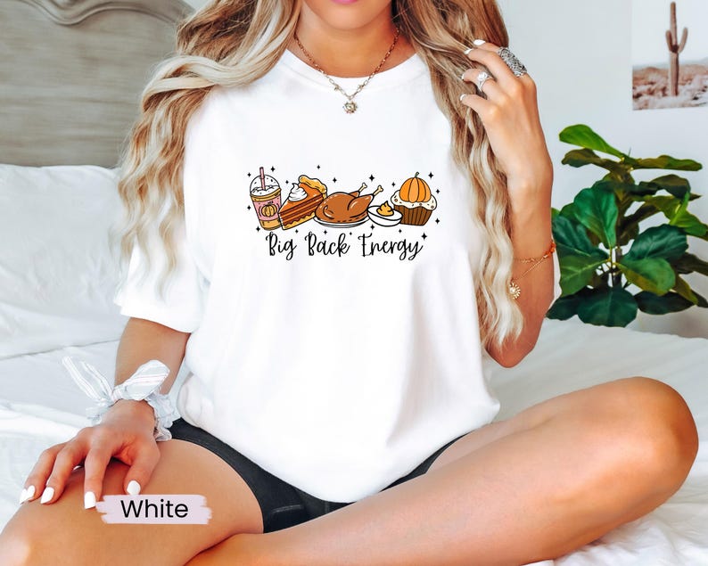 Puede incluir: Camiseta blanca con un gr&aacute;fico de comida de Acci&oacute;n de Gracias: un latte de especias de calabaza, tarta, pavo y una magdalena de calabaza. El texto "Big Pack Energy" est&aacute; escrito en negro. La camiseta es de estilo casual.