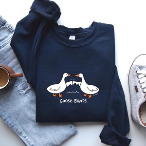 May include: A navy blue sweatshirt with a white graphic of two geese bumping into each other. The text "Goose Bumps" is printed below the geese.