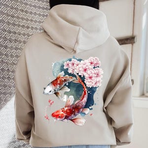 Puede incluir: Una sudadera con capucha beige con un dise&ntilde;o de acuarela de dos peces koi nadando a trav&eacute;s de flores de cerezo rosas.