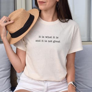May include: Cream-colored t-shirt with the text "it is what it is and it is not great" in black. A straw hat with a black band is held near the shirt. The shirt is paired with white shorts.