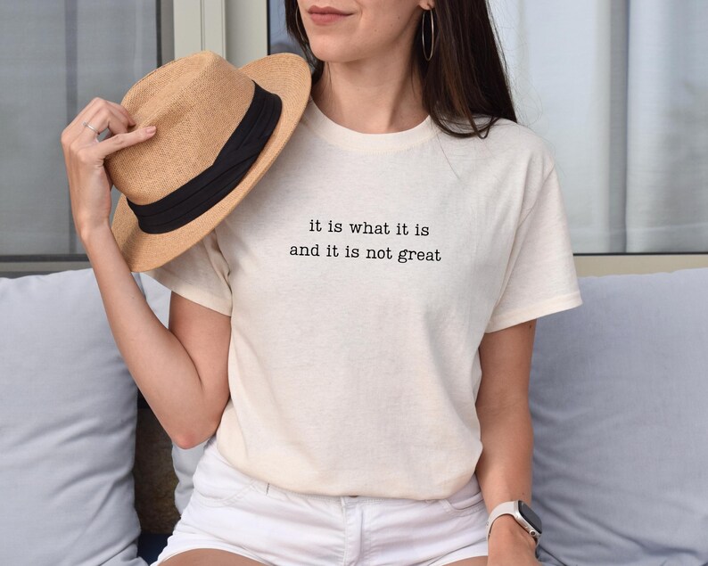 May include: Cream-colored t-shirt with the text "it is what it is and it is not great" in black. A straw hat with a black band is held near the shirt. The shirt is paired with white shorts.