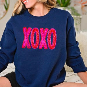 以下が含まれることがあります： ピンクのグリッターのXOXOグラフィックが施されたネイビーのトレーナー