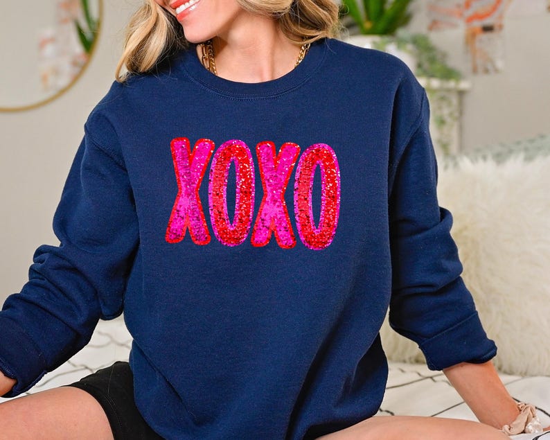 以下が含まれることがあります： ピンクのグリッターのXOXOグラフィックが施されたネイビーのトレーナー