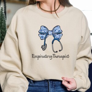 May include: A light beige sweatshirt with a blue and white floral bow and a stethoscope. The text "Respiratory Therapist" is printed below the bow.