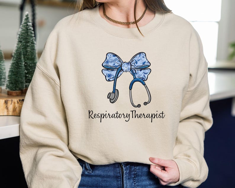 May include: A light beige sweatshirt with a blue and white floral bow and a stethoscope. The text "Respiratory Therapist" is printed below the bow.