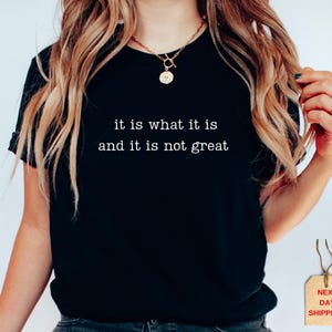 May include: Black t-shirt with the text "it is what it is and it is not great" in white. The shirt is a crew neck style and is being worn by a person. A gold necklace and rings are also visible.