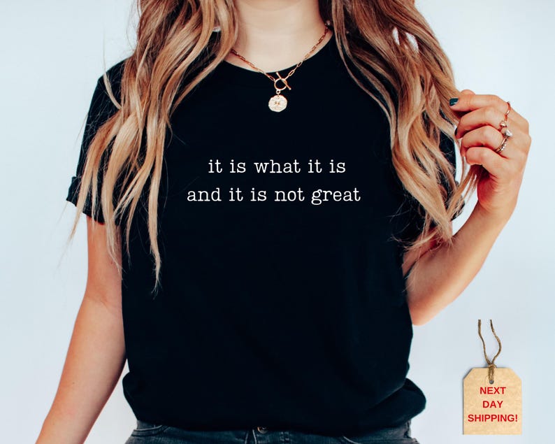 May include: Black t-shirt with the text "it is what it is and it is not great" in white. The shirt is a crew neck style and is being worn by a person. A gold necklace and rings are also visible.