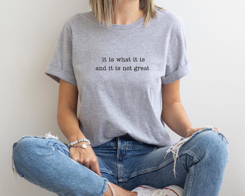 May include: A light gray t-shirt with the text "it is what it is and it is not great" in black. The shirt is worn with blue jeans. The sleeves are rolled up.