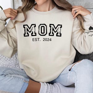 Puede incluir: Una sudadera de color crema con el texto "MOM EST. 2024" en negro. La sudadera tiene un corazón negro con el texto "Katie" en la manga izquierda.