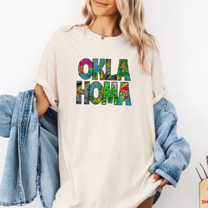 May include: Cream-colored t-shirt with the word "OKLAHOMA" in large, colorful, psychedelic-style letters. The letters are filled with a mix of vibrant colors, including green, blue, pink, and yellow. The shirt is worn with a denim jacket.