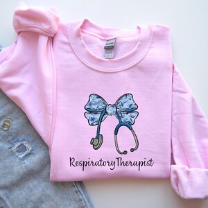 May include: Pink sweatshirt with a blue and white bow and stethoscope graphic. The text "Respiratory Therapist" is printed below the graphic.