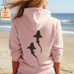 May include: Pink hoodie with a black shark silhouette graphic on the back.