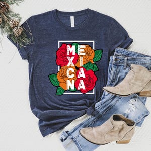 May include: A navy blue t-shirt with the word "MEXICANA" in white letters inside a white rectangle. The design includes red, orange, and yellow roses with green leaves. The shirt is paired with blue jeans and tan ankle boots.