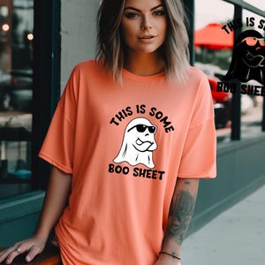 May include: A coral orange t-shirt with a white ghost wearing sunglasses and the text "This is some boo sheet".