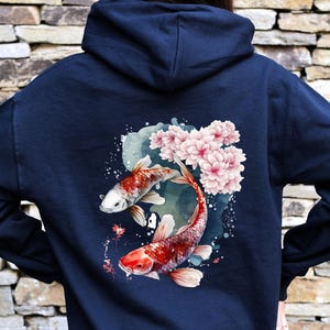 Puede incluir: Una sudadera con capucha azul marino con un dise&ntilde;o de acuarela de dos peces koi nadando entre flores de cerezo rosas.