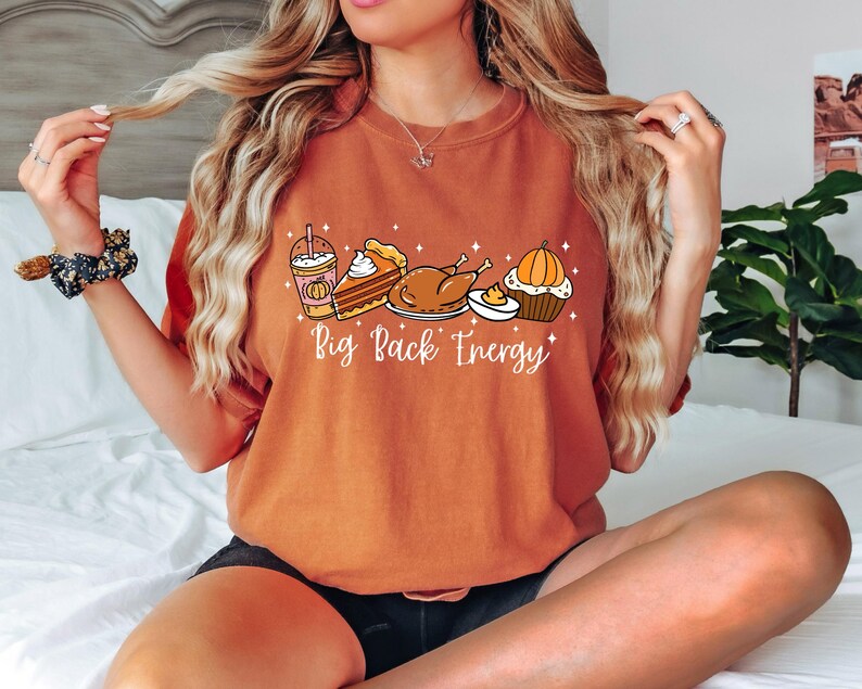Puede incluir: Camiseta naranja con un dise&ntilde;o tem&aacute;tico de Acci&oacute;n de Gracias. El dise&ntilde;o incluye un caf&eacute; con leche de calabaza especiada, tarta, pavo y una magdalena. El texto "Big Back Energy" est&aacute; impreso debajo.
