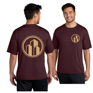May include: Maroon short-sleeve t-shirt with a gold circular logo featuring three stylized buildings. The logo is repeated on the front and back of the shirt. The shirt is worn by a person.
