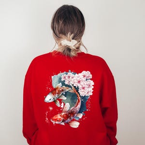 Puede incluir: Sudadera roja con un dise&ntilde;o de acuarela de dos peces koi nadando en un estanque con flores rosas.