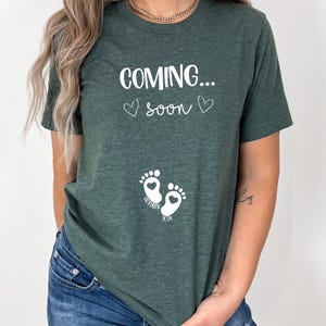 Op de afbeelding: Een donkergroen T-shirt met korte mouwen en witte tekst "COMING... soon" met twee kleine hartjes. Onder de tekst staan twee witte babyvoetjes met hartvormige centra, met de woorden "OCTOBER" en "2024" eronder gedrukt.