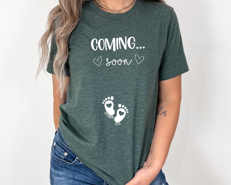 Op de afbeelding: Een donkergroen T-shirt met korte mouwen en witte tekst "COMING... soon" met twee kleine hartjes. Onder de tekst staan twee witte babyvoetjes met hartvormige centra, met de woorden "OCTOBER" en "2024" eronder gedrukt.