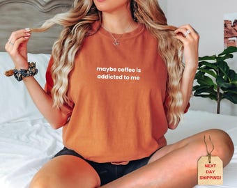 May include: A burnt orange t-shirt with the phrase "maybe coffee is addicted to me" printed in white. The shirt is a solid color and has a relaxed fit. The model is wearing black shorts.