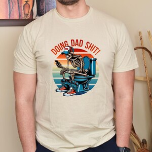 May include: Beige t-shirt with the phrase "DOING DAD SHIT!" above a skeleton graphic. The skeleton is seated on a toilet, wearing a "DAD" hat, jeans, and sneakers, and holding a phone. The design features a sunset background.