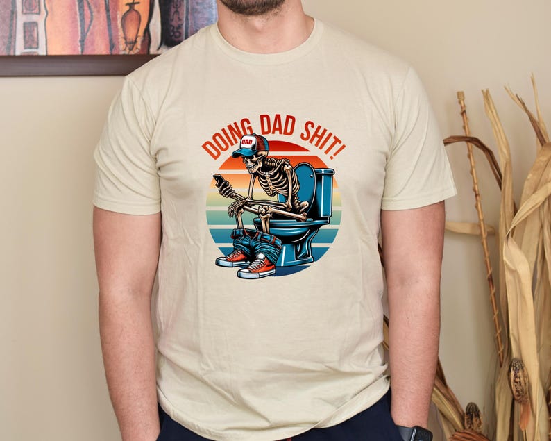 May include: Beige t-shirt with the phrase "DOING DAD SHIT!" above a skeleton graphic. The skeleton is seated on a toilet, wearing a "DAD" hat, jeans, and sneakers, and holding a phone. The design features a sunset background.