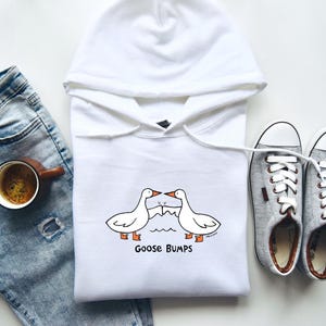 May include: White hooded sweatshirt with a graphic of two geese with the text "Goose Bumps" printed below. The sweatshirt is paired with blue jeans and gray sneakers.