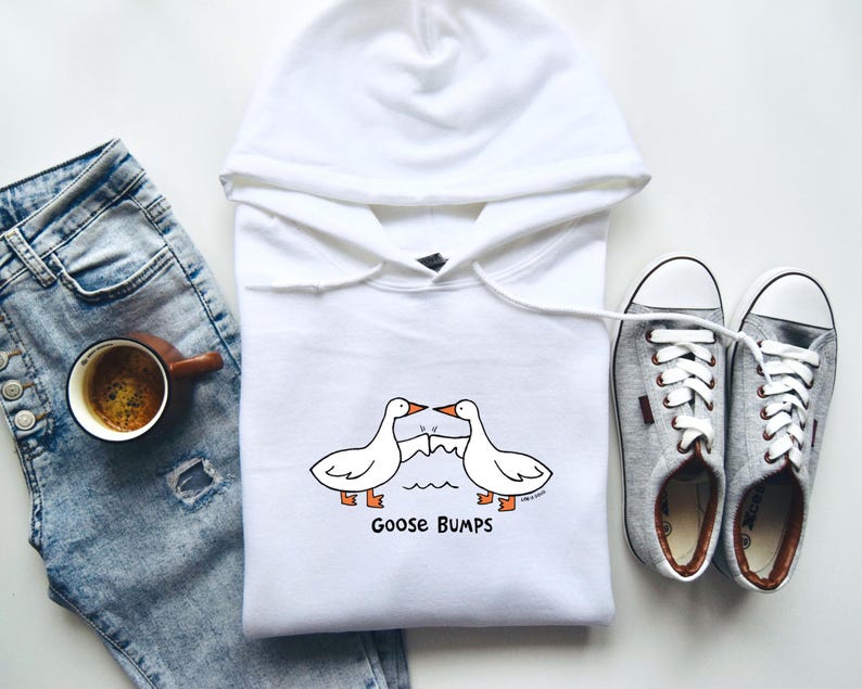 May include: White hooded sweatshirt with a graphic of two geese with the text "Goose Bumps" printed below. The sweatshirt is paired with blue jeans and gray sneakers.