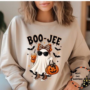 May include: A beige crewneck sweatshirt featuring a graphic of a ghost wearing sunglasses, a leopard print bow, and leopard print sneakers. The ghost holds an orange drink and a jack-o'-lantern bag, surrounded by bats and the text BOO-JEE.