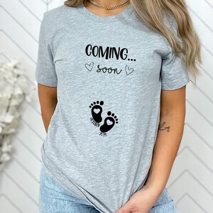 Op de afbeelding: Een lichtgrijs t-shirt met korte mouwen en de zwarte tekst "COMING..." boven "soon", geflankeerd door twee kleine hartjes. Daaronder staat een afbeelding van twee babyvoetjes met hartvormige hielen en de zwarte tekst "OCTOBER 2024".